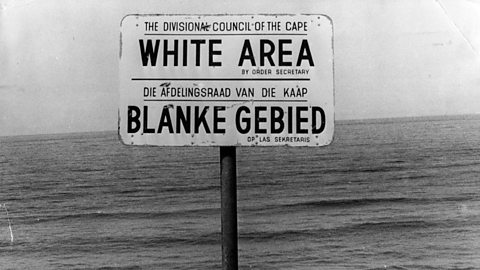 A black and white photograph of sign that says 'white area' and then the same below in Afrikaans. The sign is against the backdrop of the sea