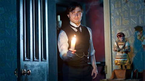 An image of the actor Daniel Radcliffe as Arthur Kipps in the 2012 film holding a candle and walking into a room with a scared expression on his face