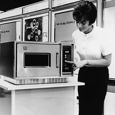 A woman wearing a white blouse and a black pencil skirts demonstrating microwave oven.
