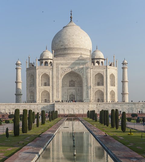 A large white building with pillars and domes stands against a background on blue sky. In the foreground, a thin man made strip of water reflects the building, grass lines the side of the water with small neat trees along it.