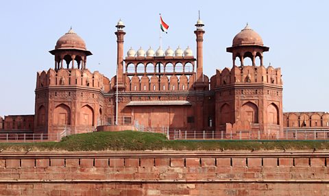 An ornate red stone building stand against a blue sky background It has two turrets, one on each side and pillars and arches between the turrets. In the foreground there is grass and another red brick wall.