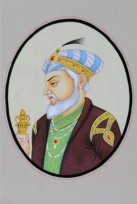 An image of a man wearing a blue and white turban, wearing a green top and burgandy coat with gold decorations - The man holds a small gold trinket.