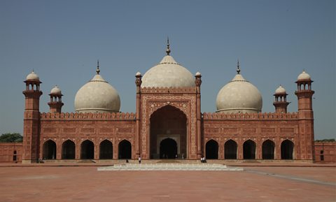 A large red brick building with arches and white domes stands against a blue sky background on a red brick foreground.