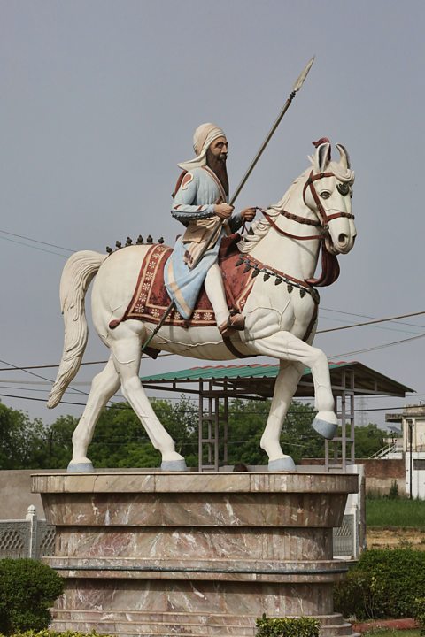 A painted statue on a plinth - The statue is of a man in blue clothes, holding a spear riding a white horse.