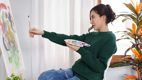 Young woman holding paintbrush in front of canvas to measure proportion.