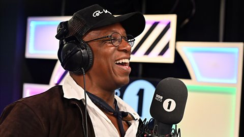 Retired footballer Ian Wright in a Radio1 studio, speaking on a radio microphone, wearing headphones and a baseball cap.