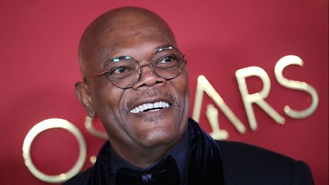 Actor Samuel L. Jackson standing in front of a crimson background with the word 'Oscars' in gold