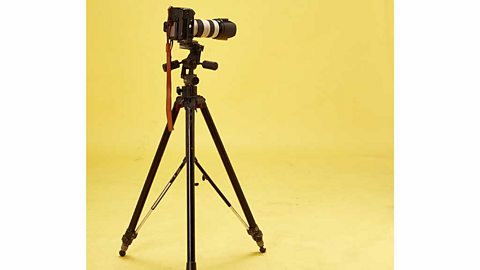 A camera with a big lens on a tripod against a yellow background