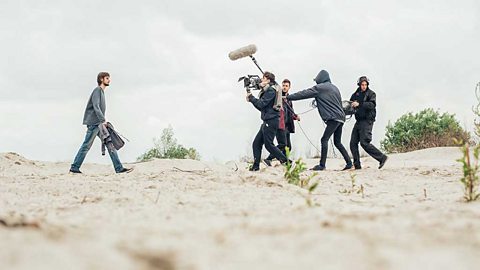 A film crew with a camera and boom mic filming a man walking on the beach