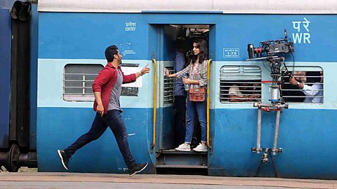 A man running up to a woman on a train. A film camera attached to the side of the train.