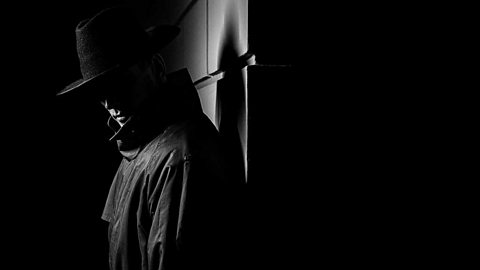 Noir style black and white image of a shadowy figure in a hat and coat