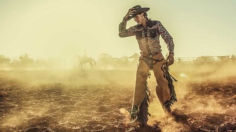 A cowboy at a dusty rodeo holding his hat. Horses in the background