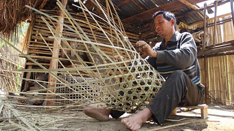 A man weaving a basket in a traditional manner in a bamboo hut