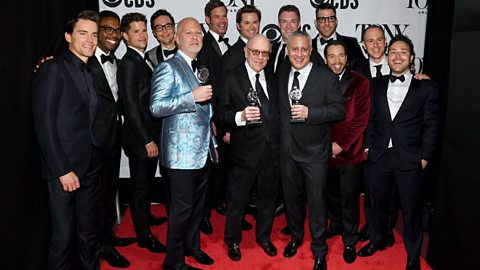 A group of men in suits stand in a group on a red carpet, the front three holding Tony Awards