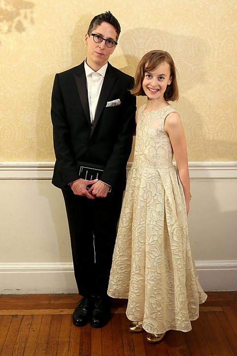 A middle aged woman with short hair and wearing a dark suit stands next to a girl wearing a long cream dress