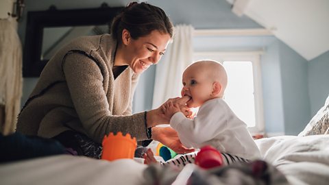 Baby girl is sitting with her mother on the bed. She is trying to teethe on her mother's finger.