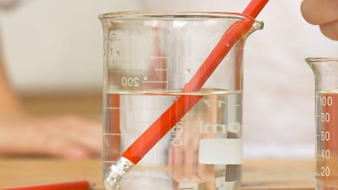 A pencil half-submerged in water, in a beaker. It looks broken at the water's edge.