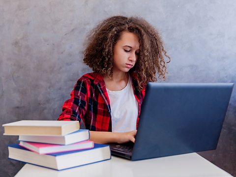 A young student sat at a desk working on their laptop with a stack of books next to them