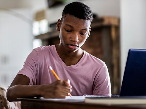 Student writing with a pencil in front of a laptop