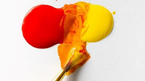 A paintbrush mixes red and yellow paint on a white surface. The paint is orange in the middle where the two colours have mixed.