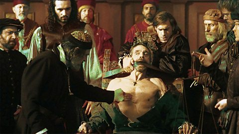 Antonio is tied to a chair with a leather belt holding him down. His shirt is pulled open and Shylock is pointing to the part of his chest from where he plans to take the flesh. Bassanio stands behind the chair with a worried expression on his face.