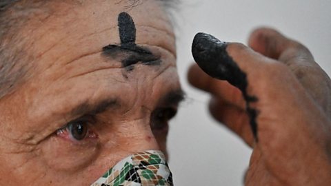 An elderly woman wearing a face mask has a dark cross drawn onto her forehead using ash. The priest's finger is covered in ash.