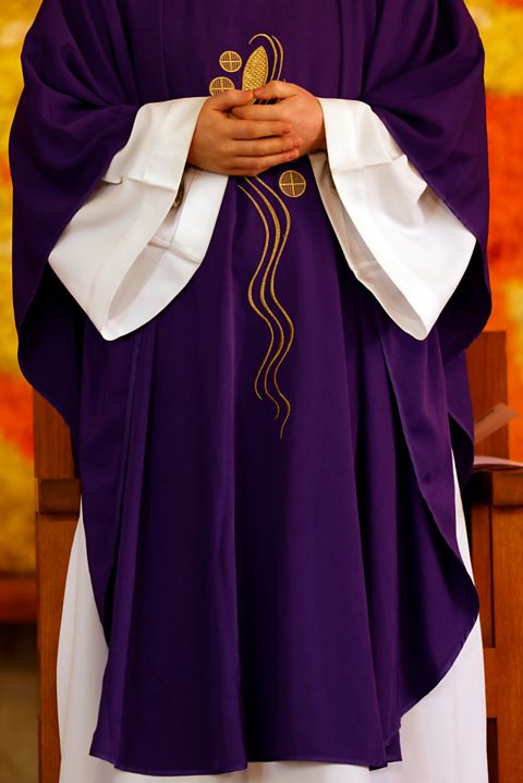 A priest wears a long purple outer robe over a longer white underlayer. The purple robe has gold detailing.