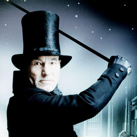 Patrick Stewart playing Scrooge in the 1999 film version. He us wearing a black coat, black gloves, tall black top hat and is raising a cane as though about to strike.