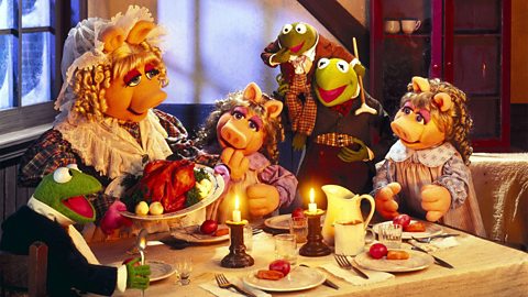 The Cratchit family from the 1992 film 'The Muppet Christmas Carol'. Miss Piggy and Kermit the Frog sit at a dinner table. Miss Piggy is holding a small, cooked turkey. They are joined by two young, female pigs and two young, male frogs. The smaller frog, Tiny Tim, is on his brother's shoulders.