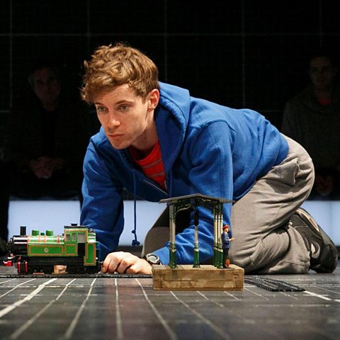 Christopher in a blue hoodie and red t-shirt. He is kneeling on the floor, playing seriously with a green toy train.