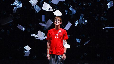 A teenage boy stands in a red t-shirt with the mathematical Pi symbol on it. In the air around him float white pieces of paper.