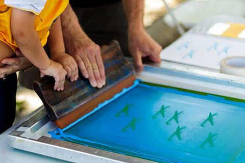 Some people screen printing. They are pushing paint over a design with a squeegee.