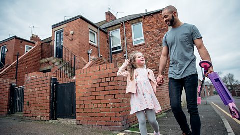 A dad walking with his daughter, he is holding a scooter.