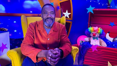 Lenny Henry wearing an orange blazer sat in a yellow wingback chair on the CBeebies Bedtime Story set.