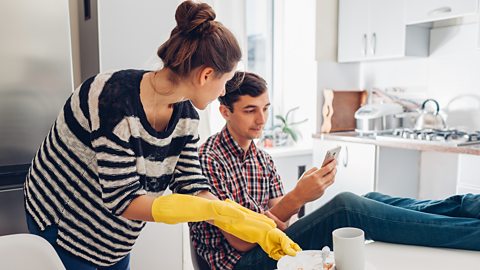 One person sits at the table with their legs on it looking at their phone while another looks angry while tidying up 