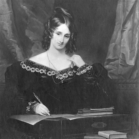 A black and white picture of Mary Shelley. Her hair is in ringlets and she is wearing a dark dress with flowers around the neckline. She is posed as though writing with a quill.