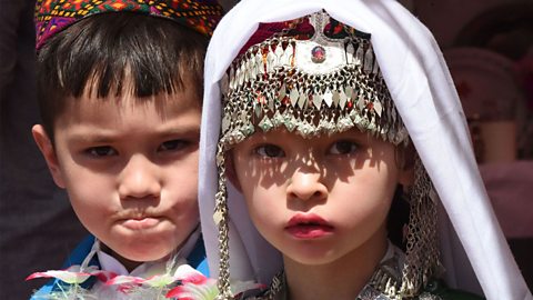 A young boy and a young girl wearing traditional Hazara dress. The boy is wearing a brightly coloured hat. The girl is wearing a heavily decorated headdress with beads and tassels. 