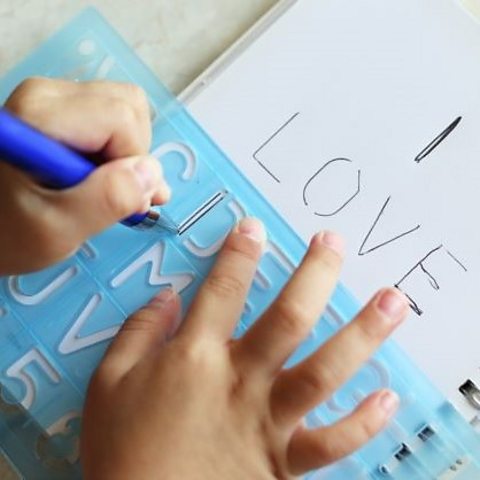 A child's hands using a pen to draw the words "I love" using alphabet stencils