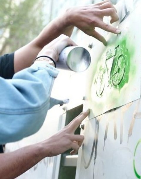 Hands seen spraying green paint onto a stencil to make a green bicycle print on a wall