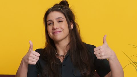 Louise Young smiling with two thumbs up in front of a yellow wall, she is wearing a black shirt.