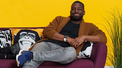 Inel Tomlinson sitting in a relaxed way on a maroon sofa in front of a yellow wall. There are cushions on the sofa including one with a picture of a smiling chimpanzee on it.