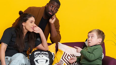 Lenny Rush, Louise Young and Inel Tomlinson on or near a maroon sofa in front of a yellow wall. There are cushions on the sofa including one with a smiling chimpanzee on it. Lenny is looking angry and pointing at Louise and Inel who are looking at Lenny with confusion on their faces.