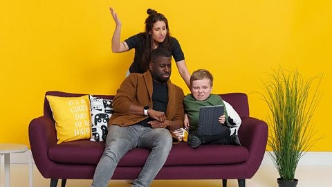 Lenny Rush, Inel Tomlinson and Louise Young sitting on or standing near a maroon sofa with a green grass plant near it. They are in front of a yellow wall. Louise has one hand in the air and looks confused, Lenny is holding a tablet and both Inel and Lenny look disgusted.