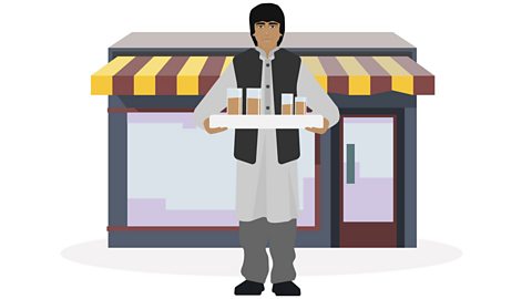 Enaiat standing in front of a shop. He is holding a tray with glasses containing tea balanced on it. 