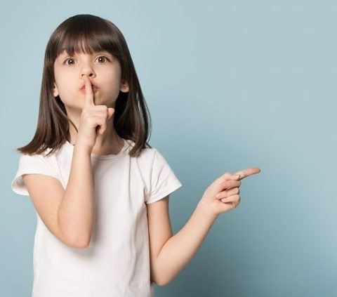 A child puts their fingers across their lips to say shush and points to their left.