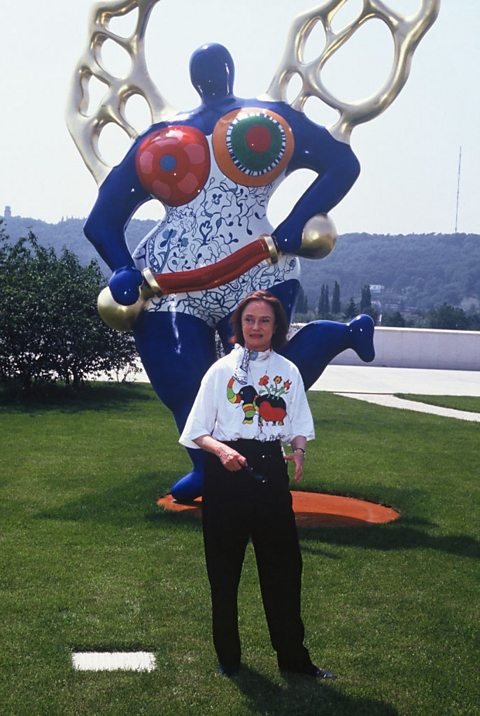 Niki de Saint Phalle stands in front of one of her Nana artworks. She is outside.