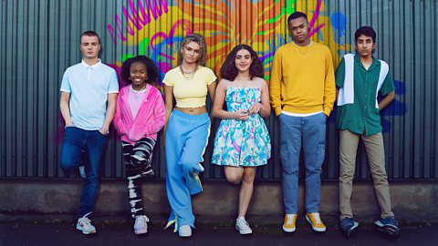 The main cast of the show Phoenix Rise standing in front of a colouful mural on a corrugated wall. 