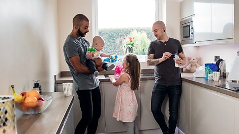 2 dads with their toddler daughter and baby in the kitchen.