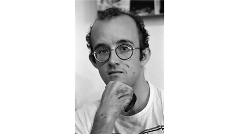 A black and white portrait of Keith Haring. He has paint on his hand and a bit on his face. He is wearing round glasses.