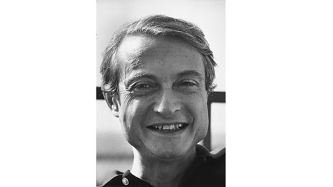 A black and white photograph of Roy Lichtenstein. He has short hair and is smiling.